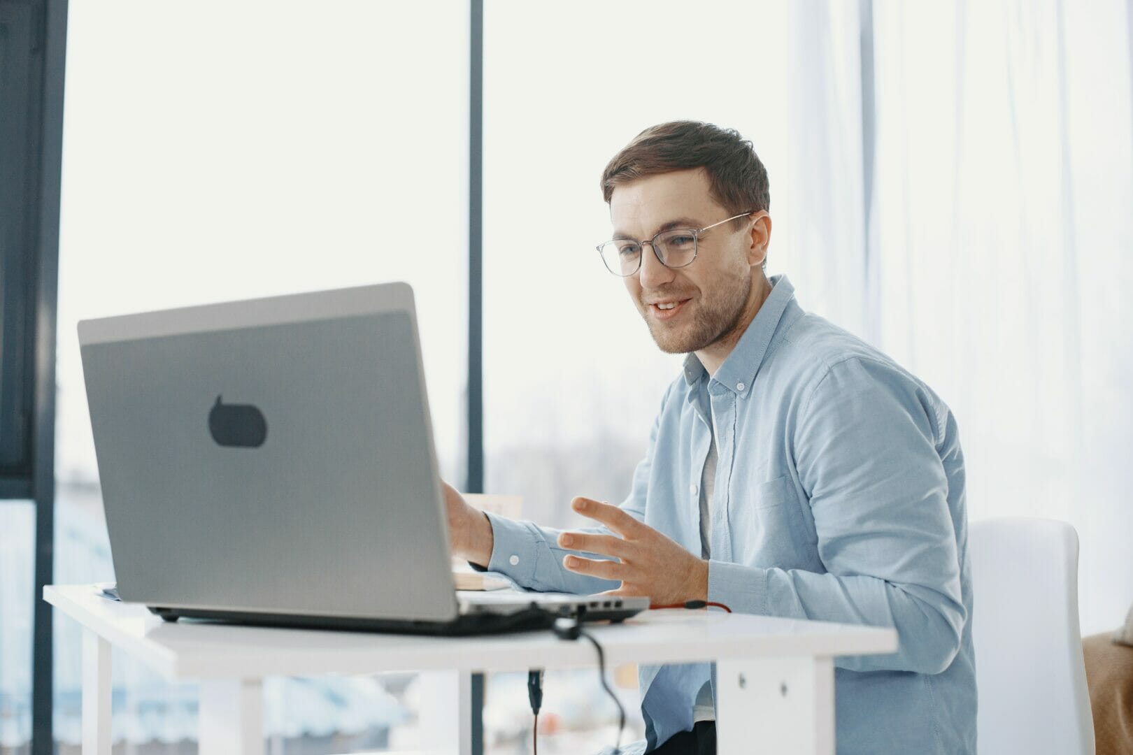 Person in glasses and a light blue sweater sitting at a desk with a laptop, gesturing as if explaining something.