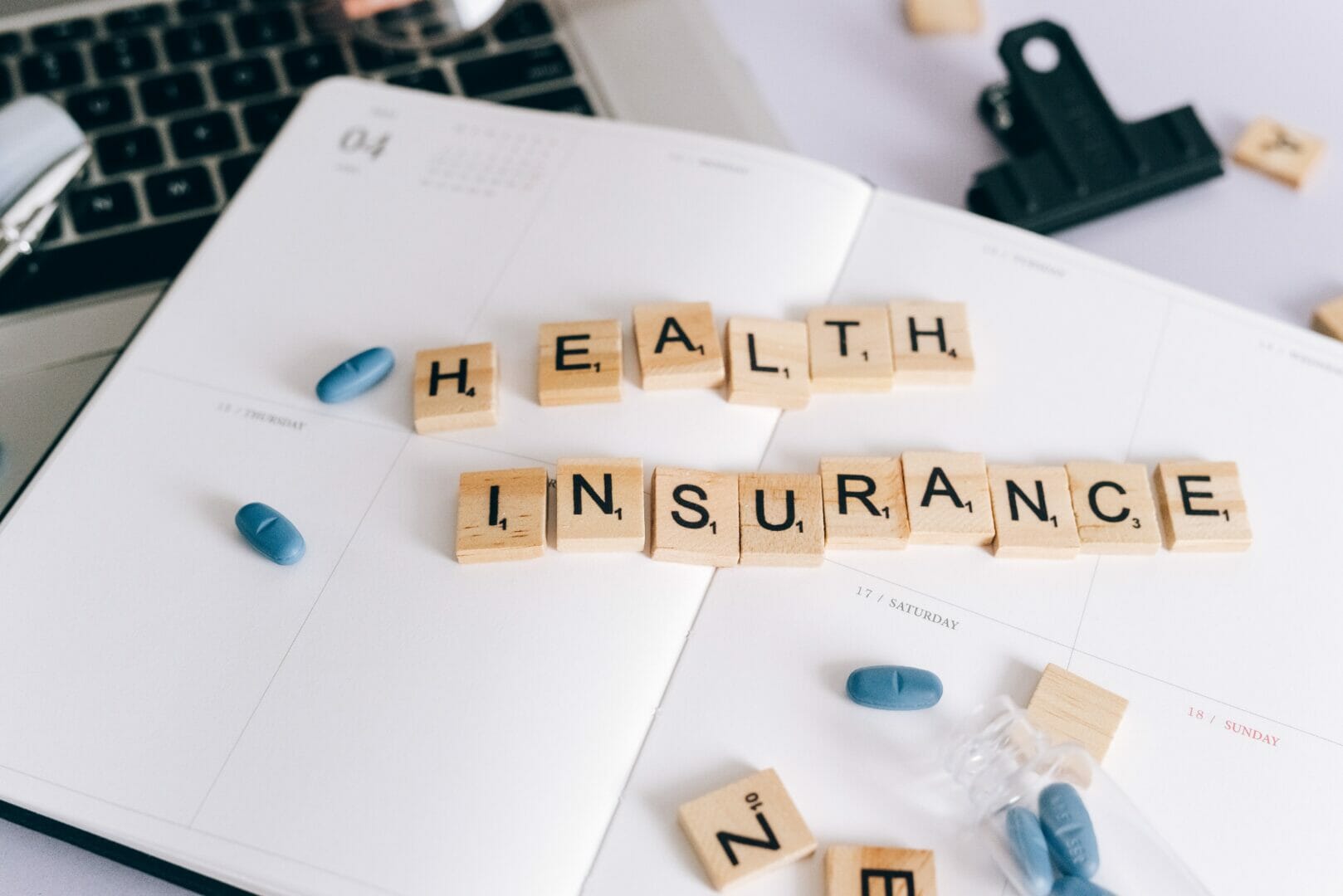 Scrabble tiles spelling HEALTH INSURANCE on a planner with pills around