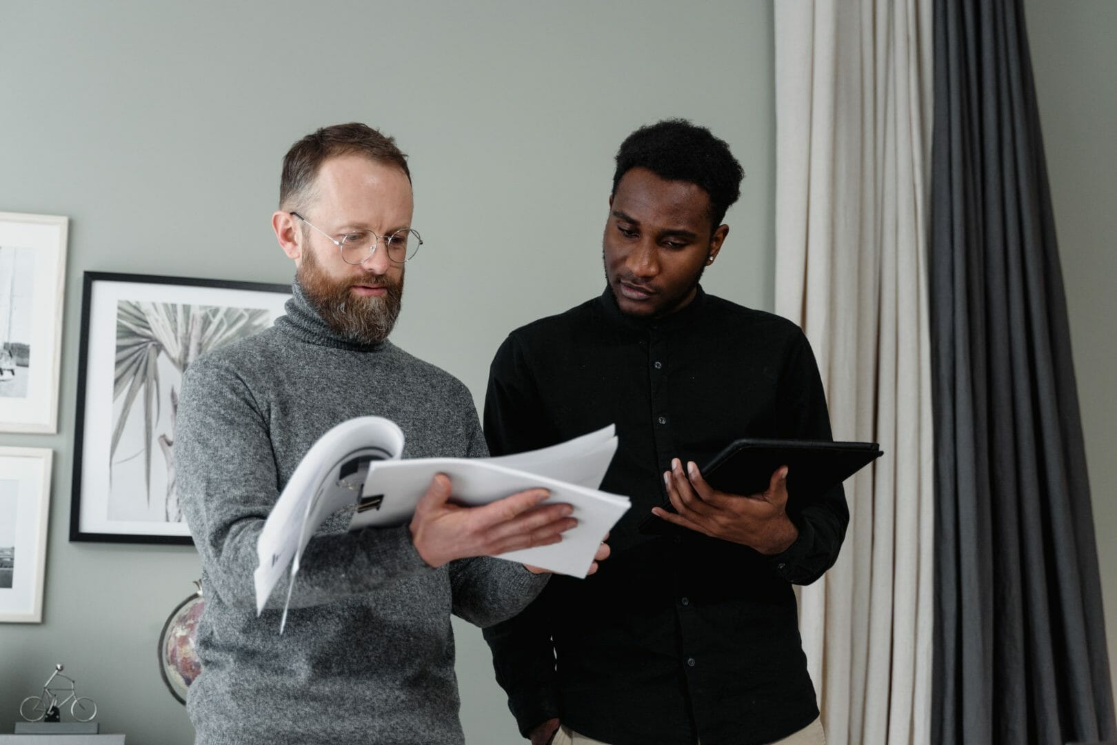 Two men in a professional setting, one with a tablet and the other with a document, discussing
