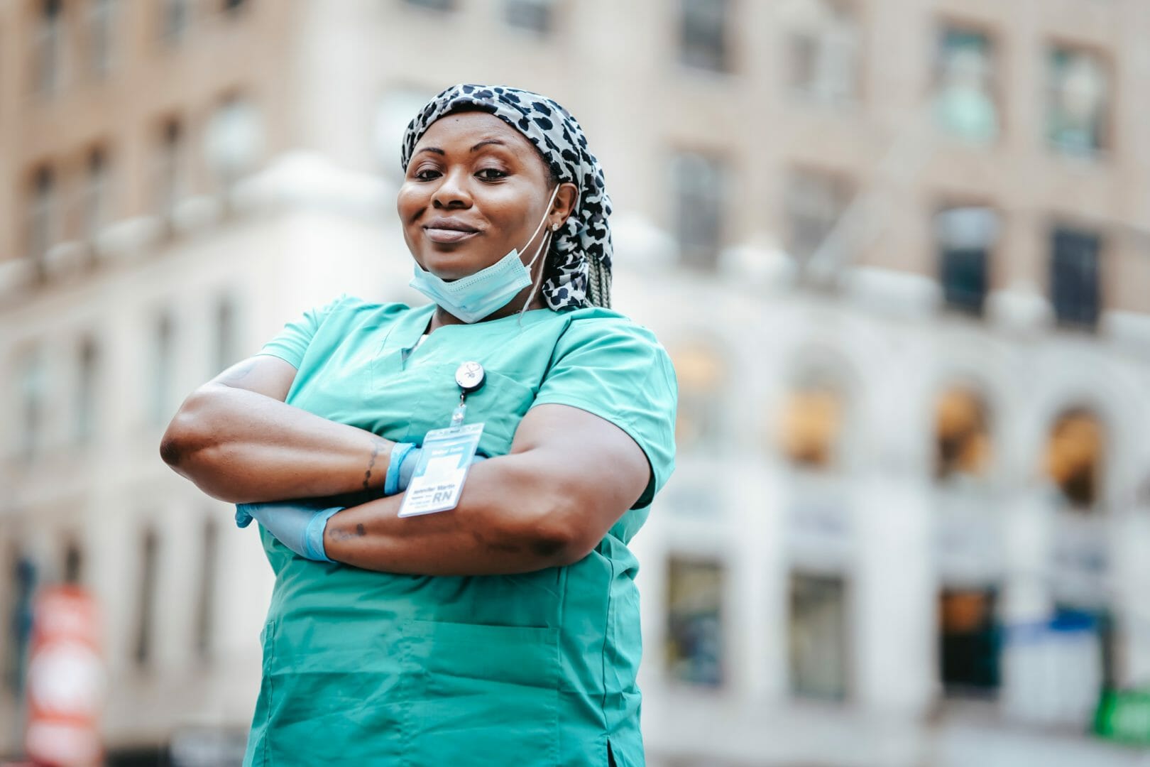 Medical professional in scrubs and mask standing with arms crossed in an urban setting
