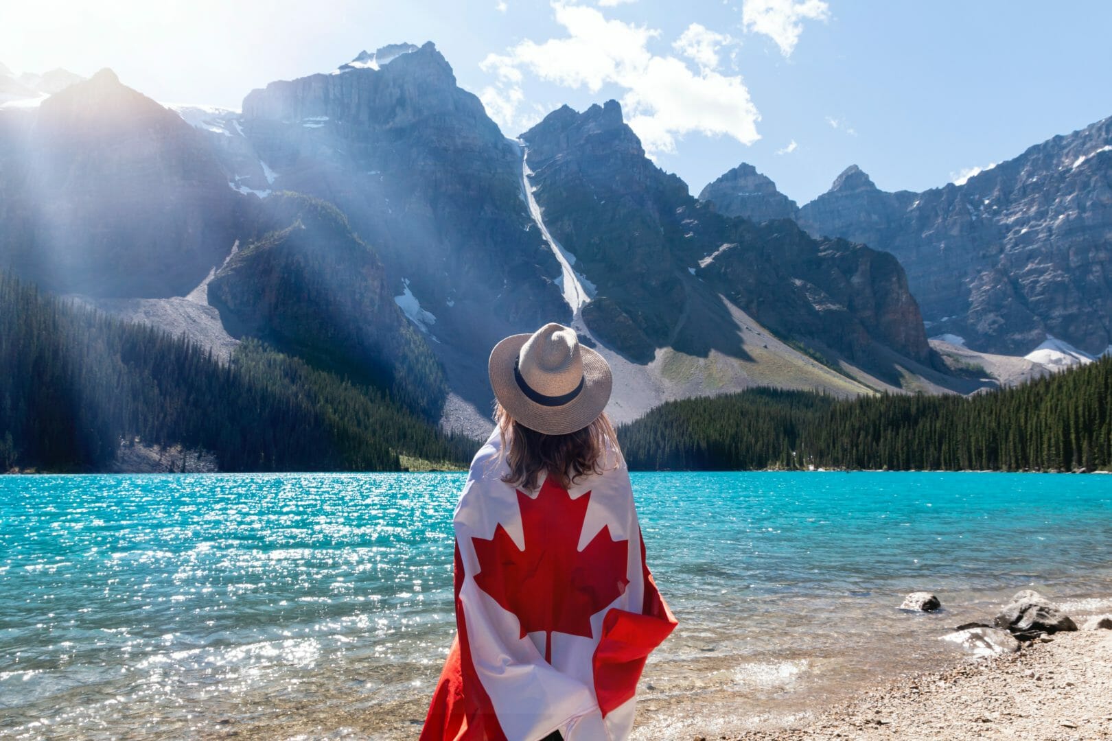 |Person standing by a lake wrapped in a Canadian flag with mountains in the background