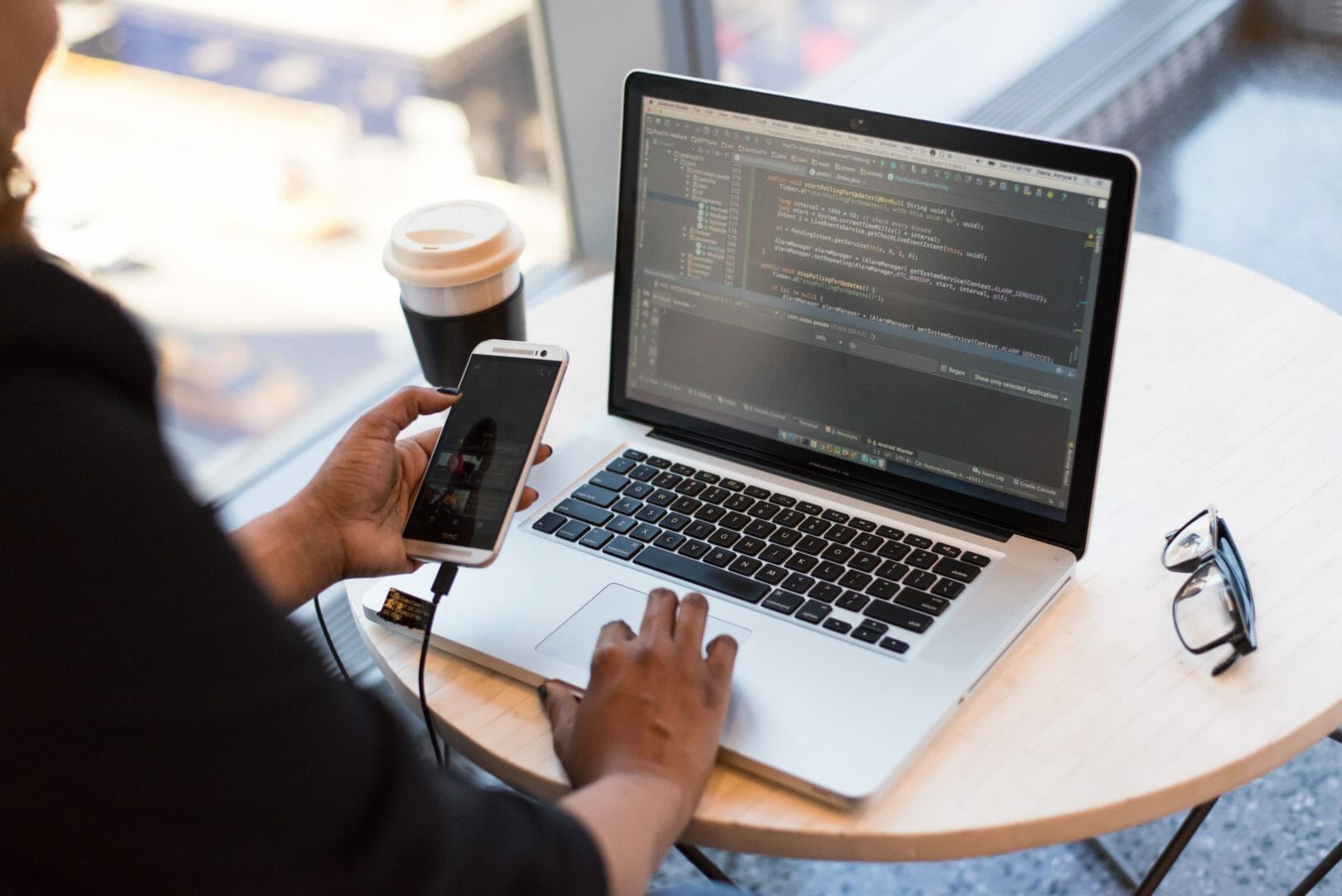 Person working on a laptop with coding software open, holding a smartphone