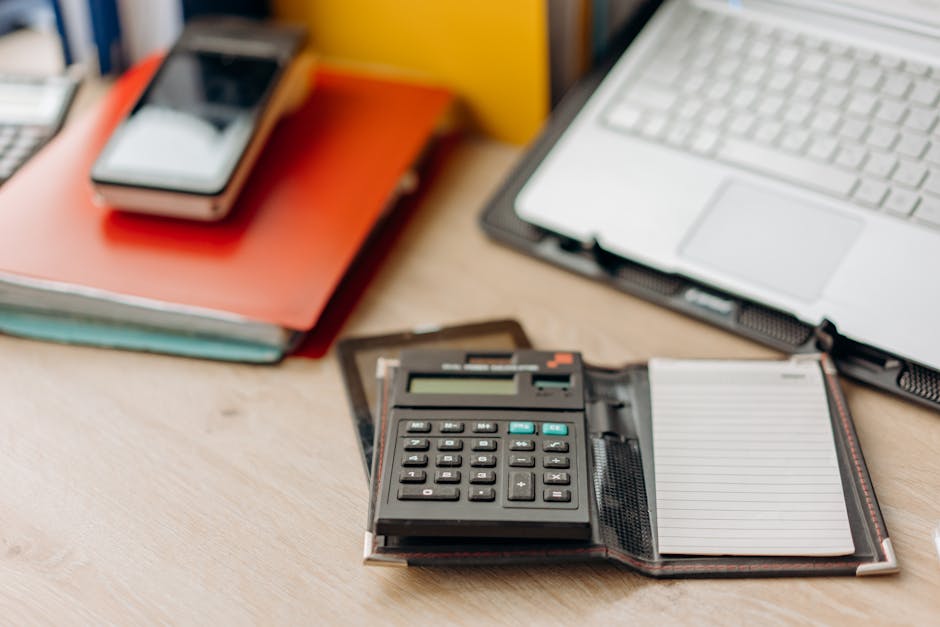Workspace with a calculator, leather notepad, smartphone on red folders, and a laptop on a wooden desk