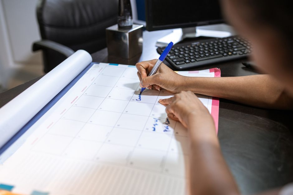 A person writing on a monthly desk calendar with a blue pen at an office desk beside a keyboard
