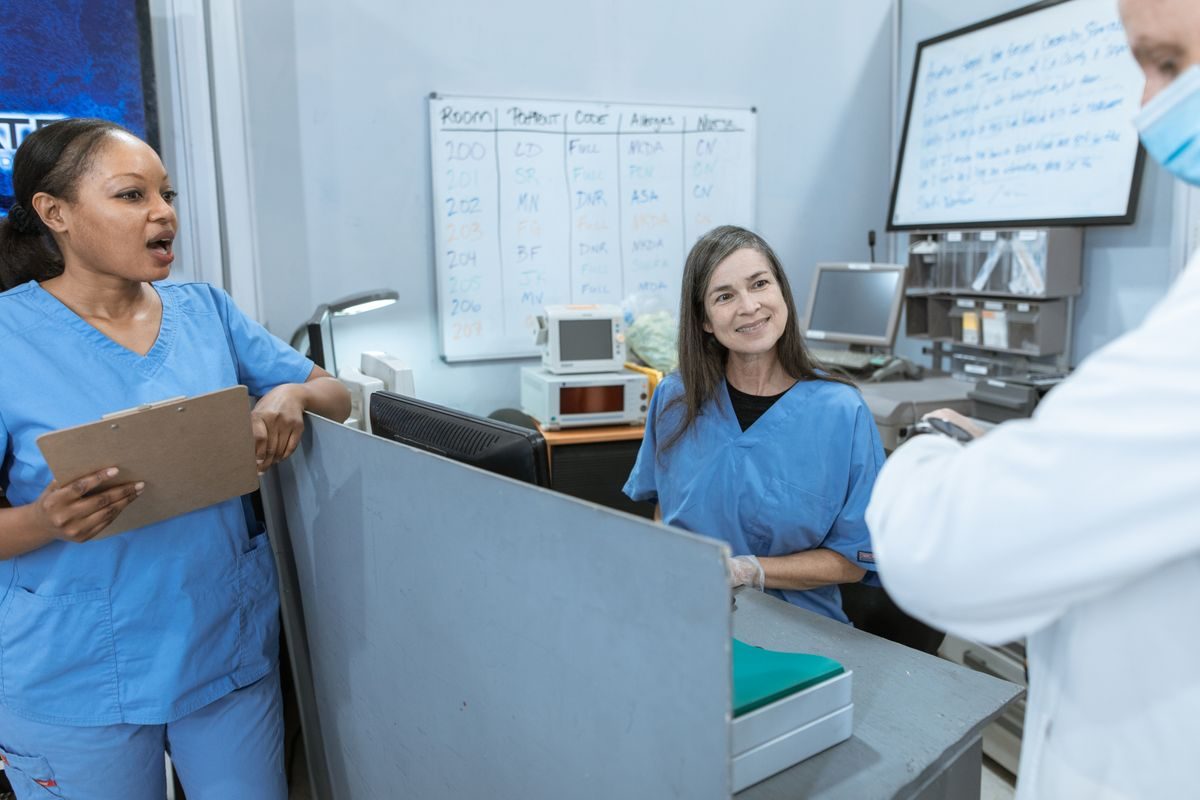 Two nurses in blue scrubs at a hospital nurses' station with a patient assignment whiteboard