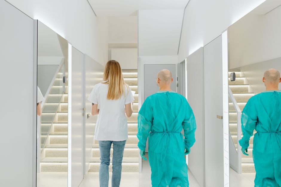 Two healthcare workers, one in surgical scrubs, walking down a bright clinic corridor toward a staircase