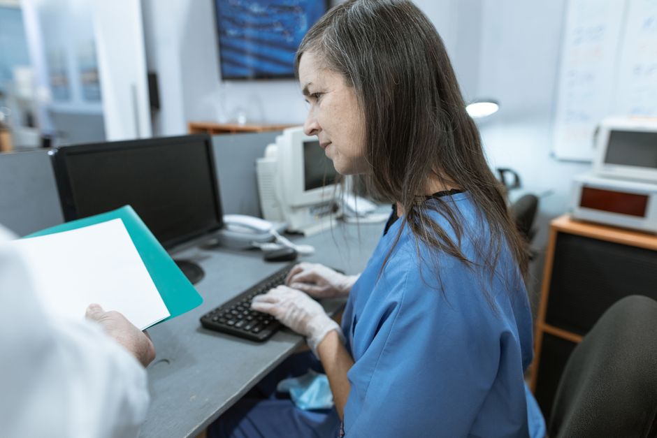 Nurse at a computer workstation in scrubs while a colleague hands over a folder of paperwork