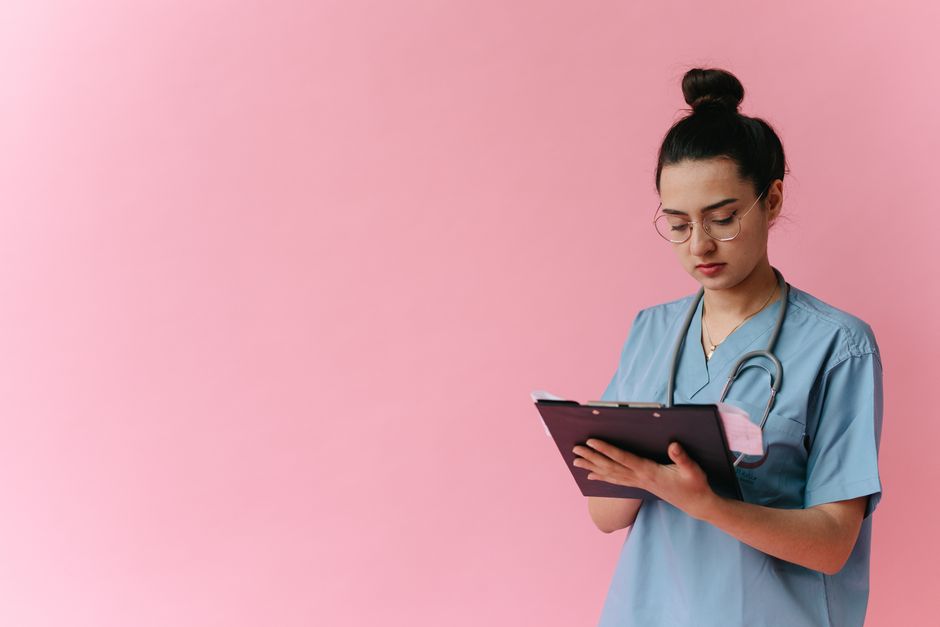 Nurse in blue scrubs reviewing notes on a clipboard, glasses, studio portrait