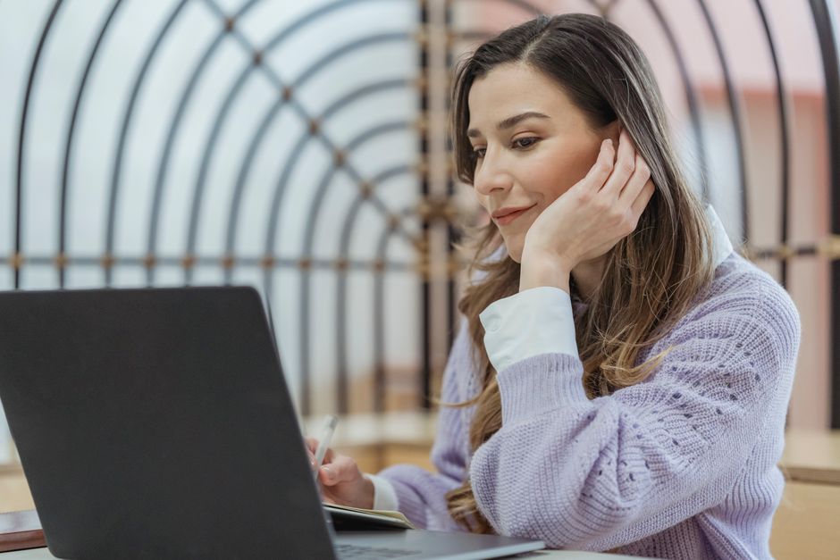 Woman in a lavender sweater smiling while resting her chin on her hand at her laptop