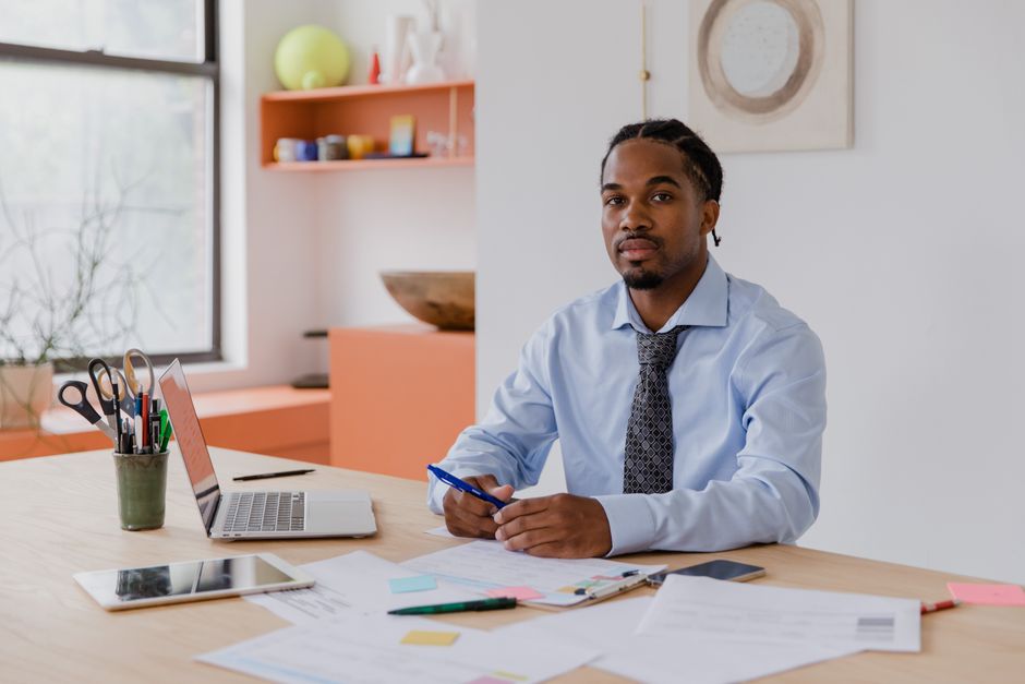 Man in a shirt and tie at a desk reviewing paperwork beside a laptop and tablet