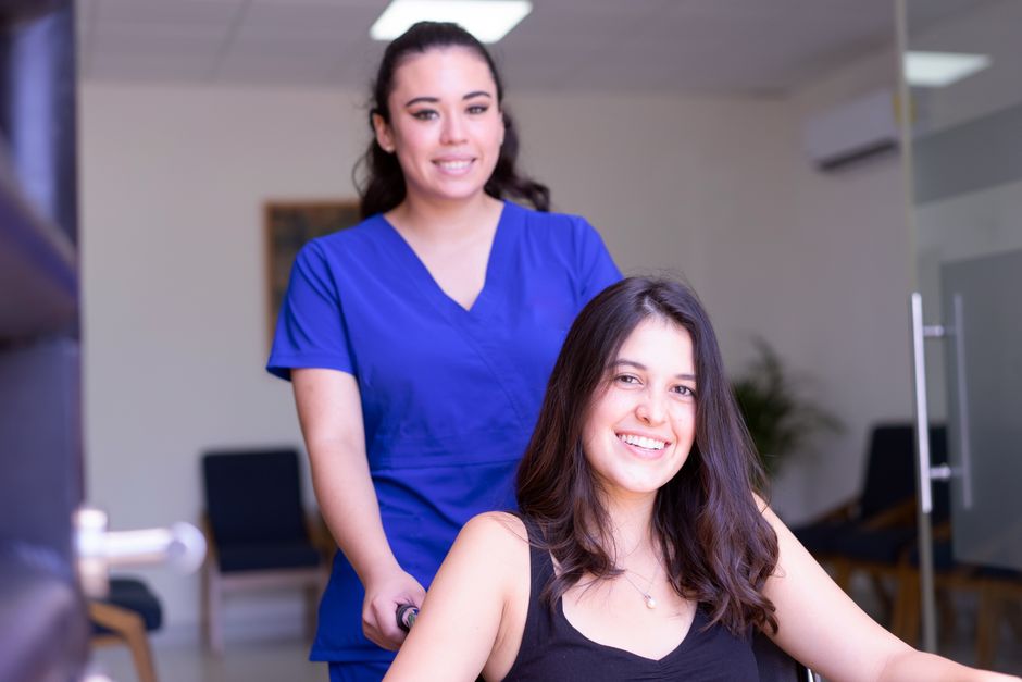 Caregiver in blue scrubs smiling while pushing a patient in a wheelchair through a clinic hallway