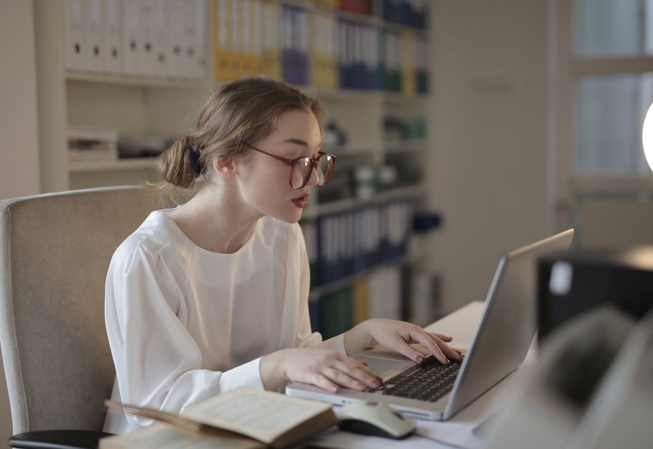 Office professional in glasses typing on a laptop at a desk with reference binders on a shelf behind her