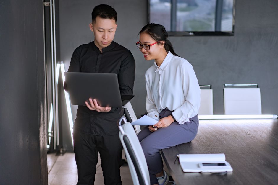 Two colleagues standing at a conference table, reviewing a laptop screen together in a modern office