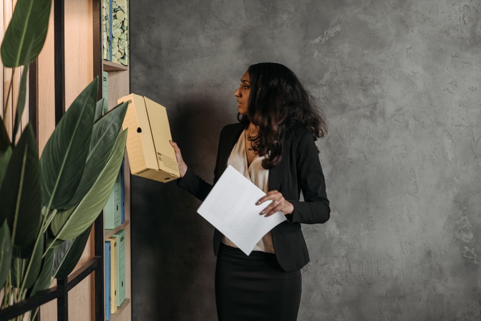 Woman in a blazer pulling a document archive box from an office file shelf while holding a sheet of paper
