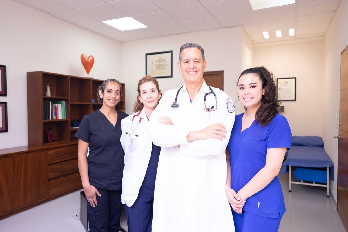 Four healthcare professionals in scrubs and white coats standing together in a clinic exam room