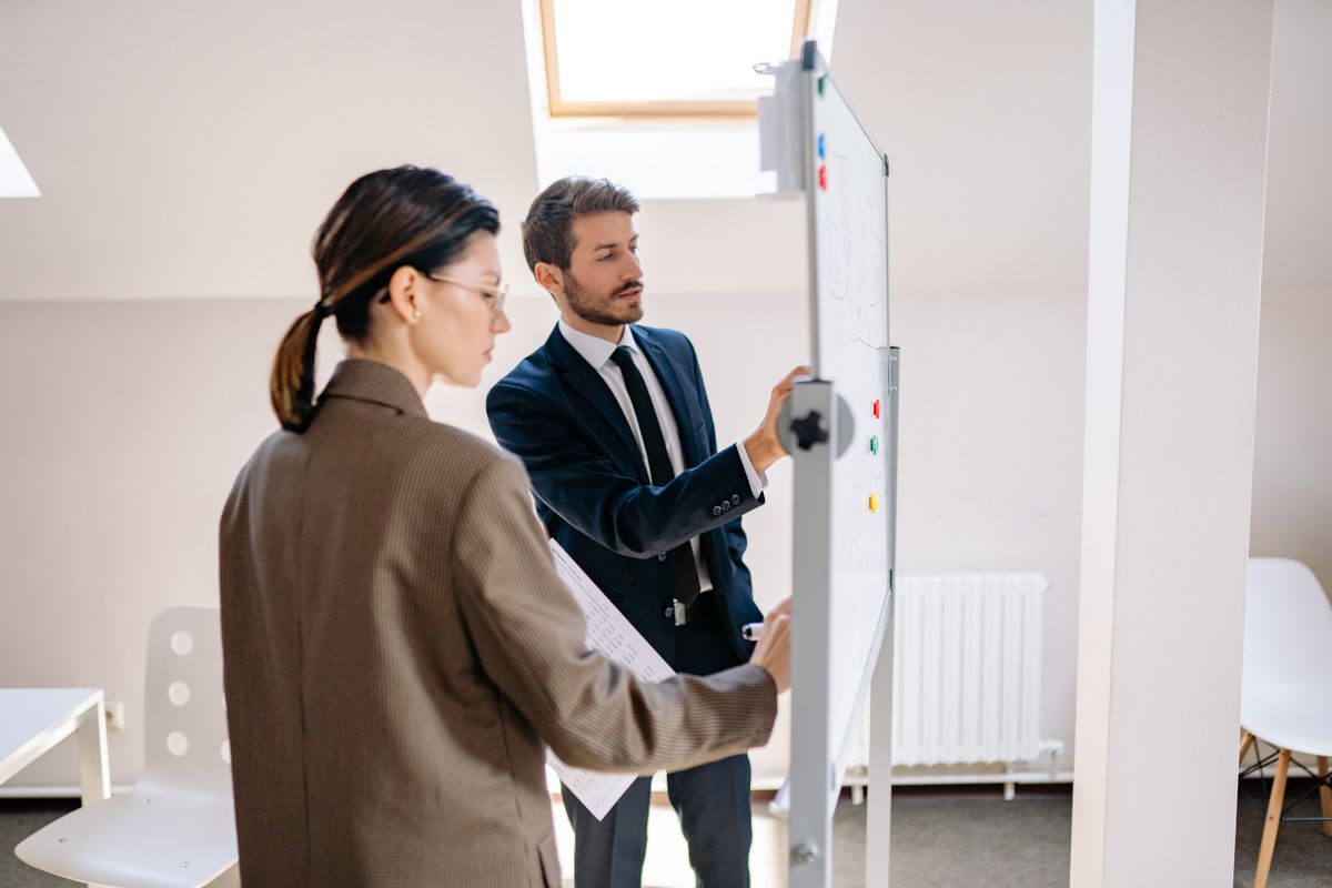 Two colleagues in business attire reviewing notes on a whiteboard, one holding printed pages, in a bright office