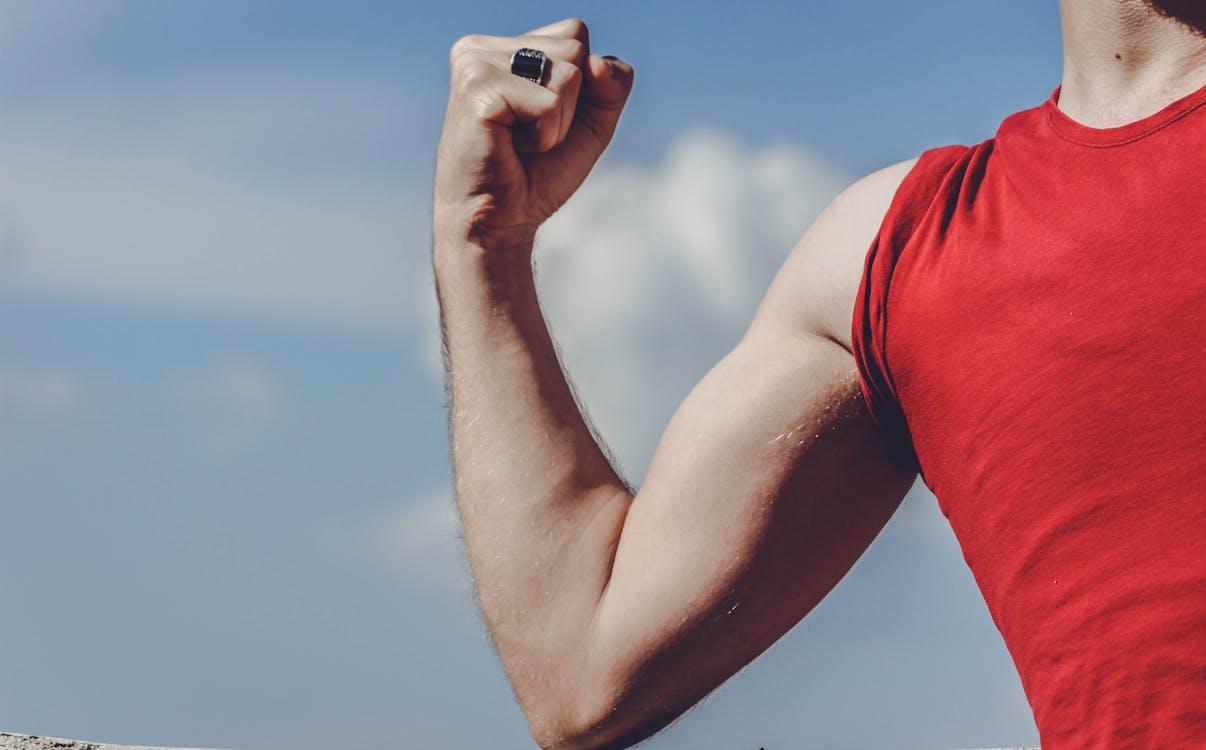 Free Close-up of a flexed arm in a red shirt against a clear blue sky, symbolizing strength and empowerment. Stock Photo