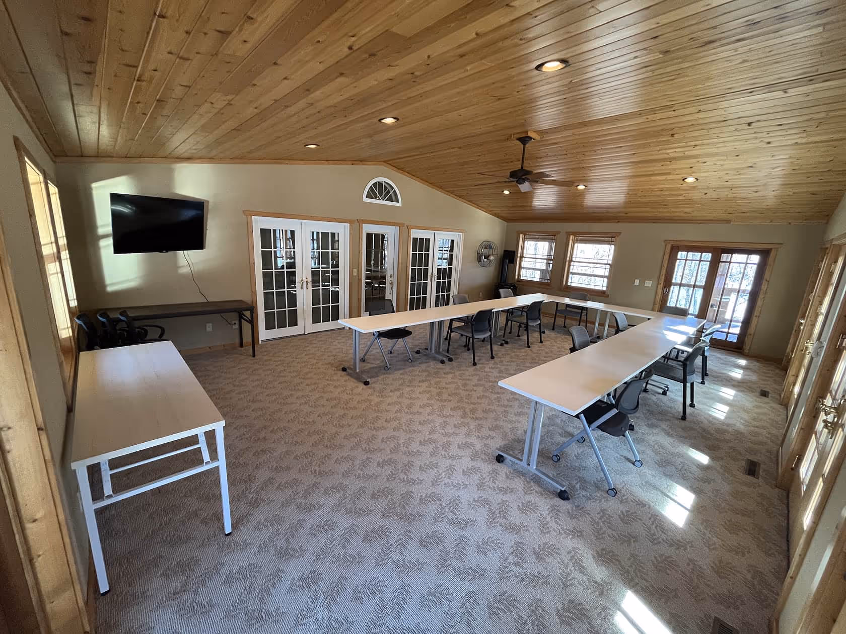 Large craft room with folding tables, chairs, and a wall-mounted TV, featuring warm wood ceilings and windows letting in natural light.