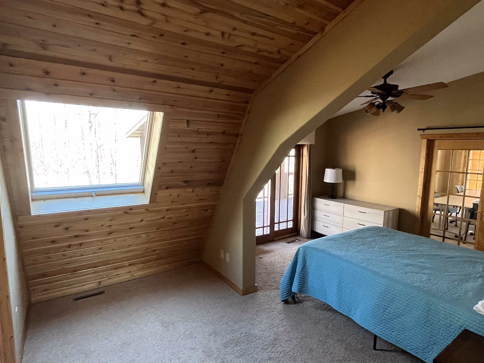 Upstairs bedroom with angled wooden ceiling, skylight window, and a blue quilt on the bed overlooking the trees.