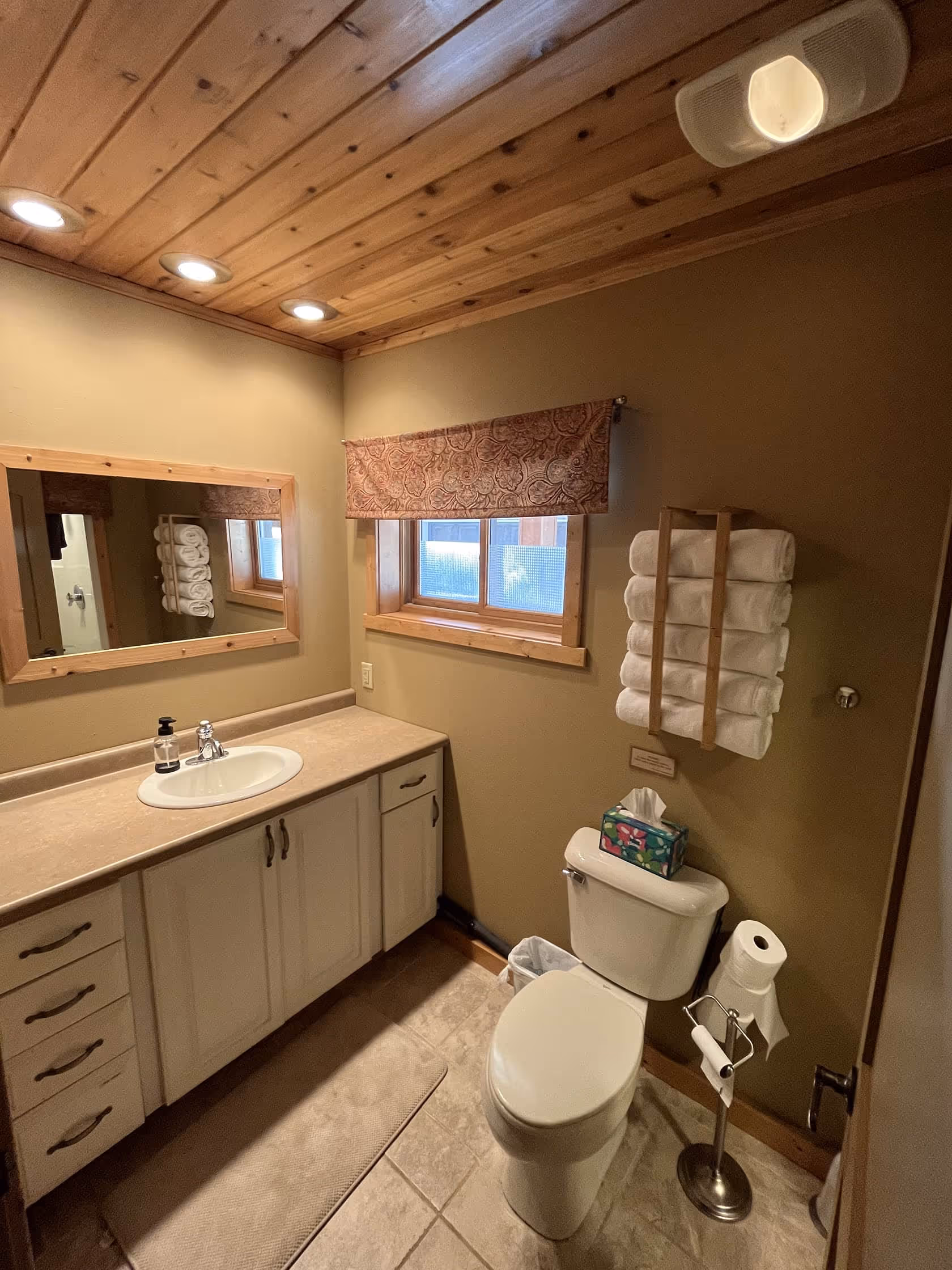 Bathroom with wood-paneled ceiling, large mirror, and neutral tones, featuring a sink, toilet, and towel rack.