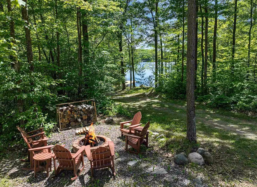 Fire pit surrounded by Adirondack chairs, with stacked wood and a clear view of Matson Lake through the trees.