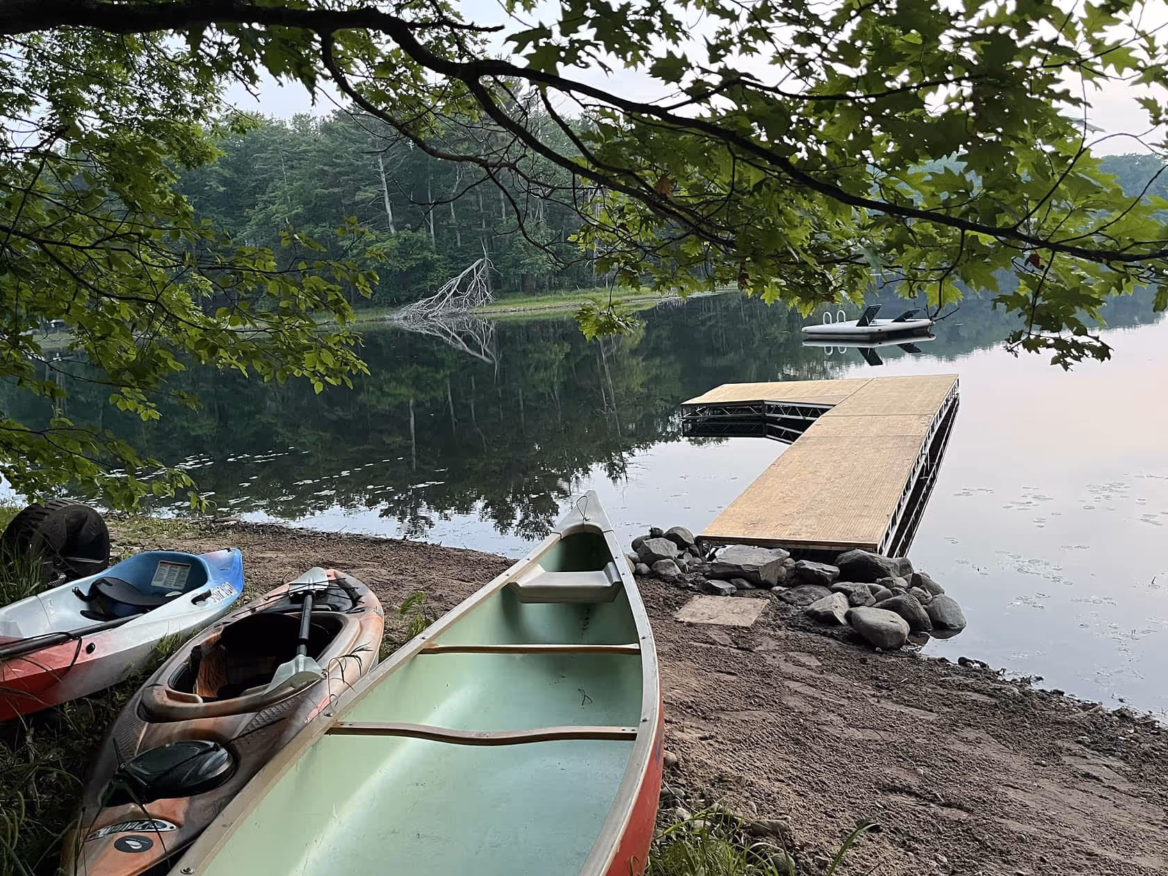 Canoes and kayaks resting on the shore beside a wooden dock on Matson Lake, surrounded by calm water and lush trees.