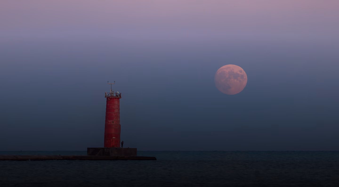 Red lighthouse on a pier at dusk with a large full moon in a clear sky.