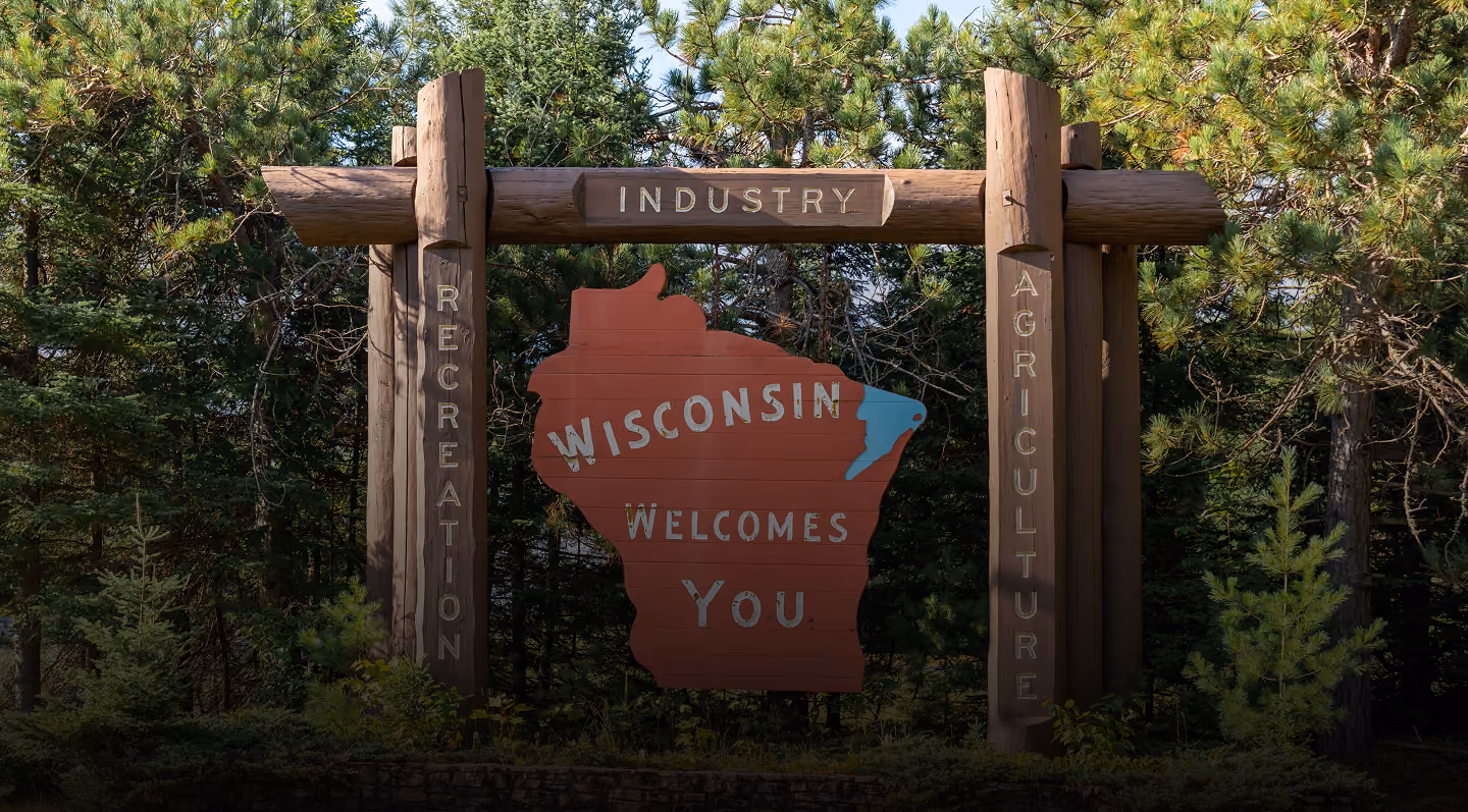 Wooden welcome sign shaped like Wisconsin with words Industry, Recreation, Agriculture, surrounded by green trees.