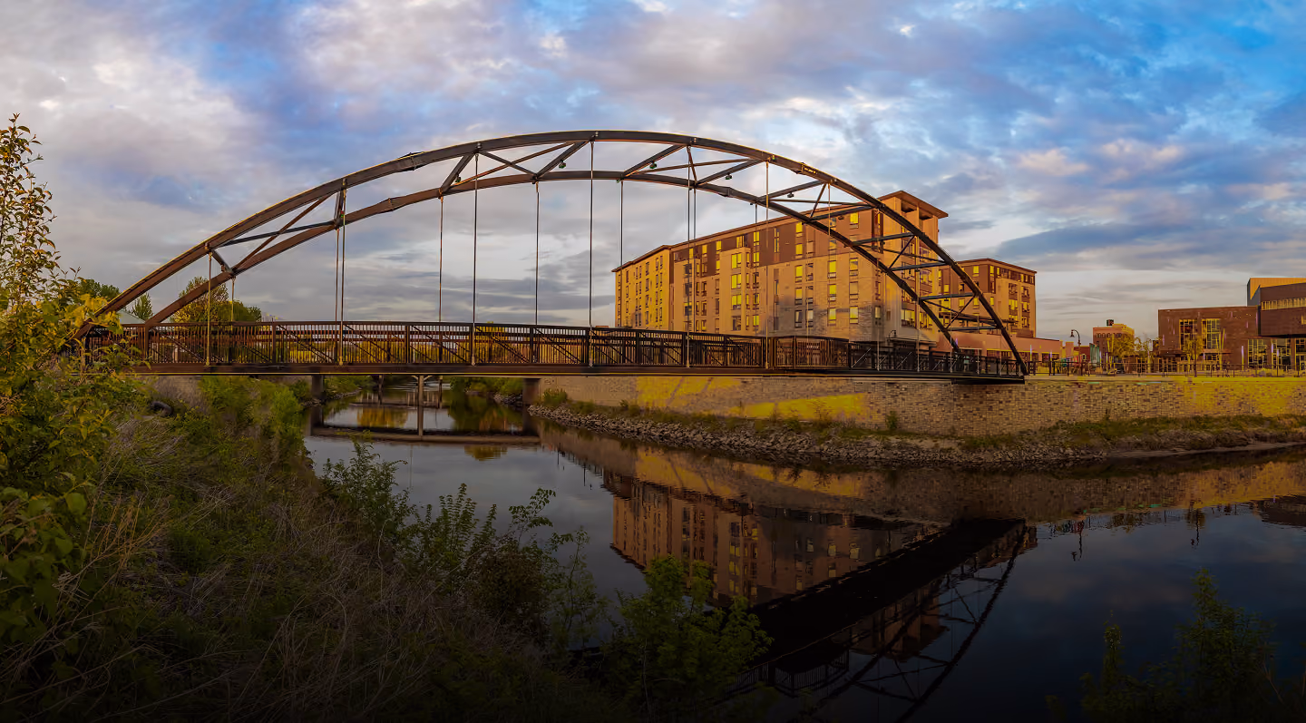 Arched pedestrian bridge over calm river reflecting nearby buildings and cloudy sky at sunset.
