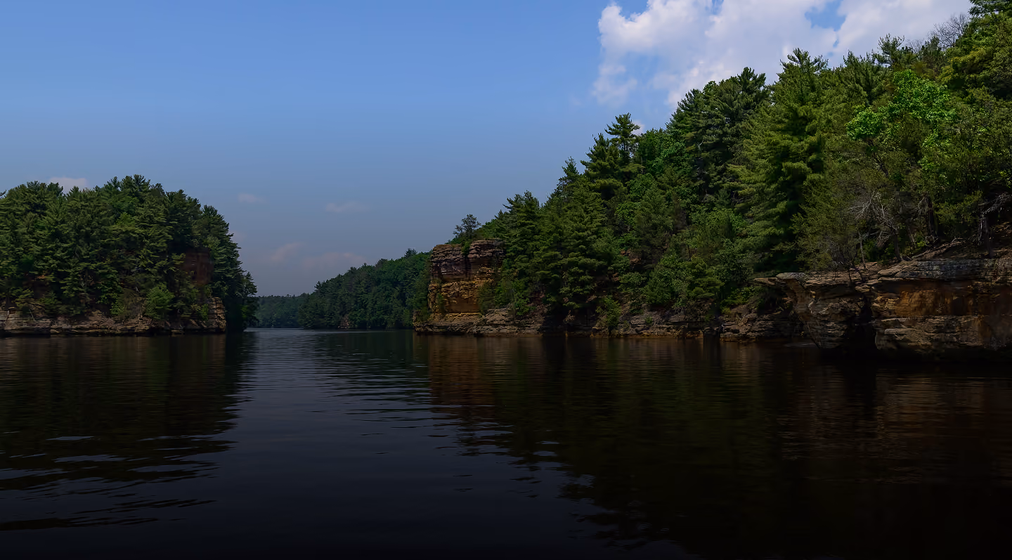 Calm river flanked by rocky cliffs and dense pine trees under a partly cloudy blue sky.