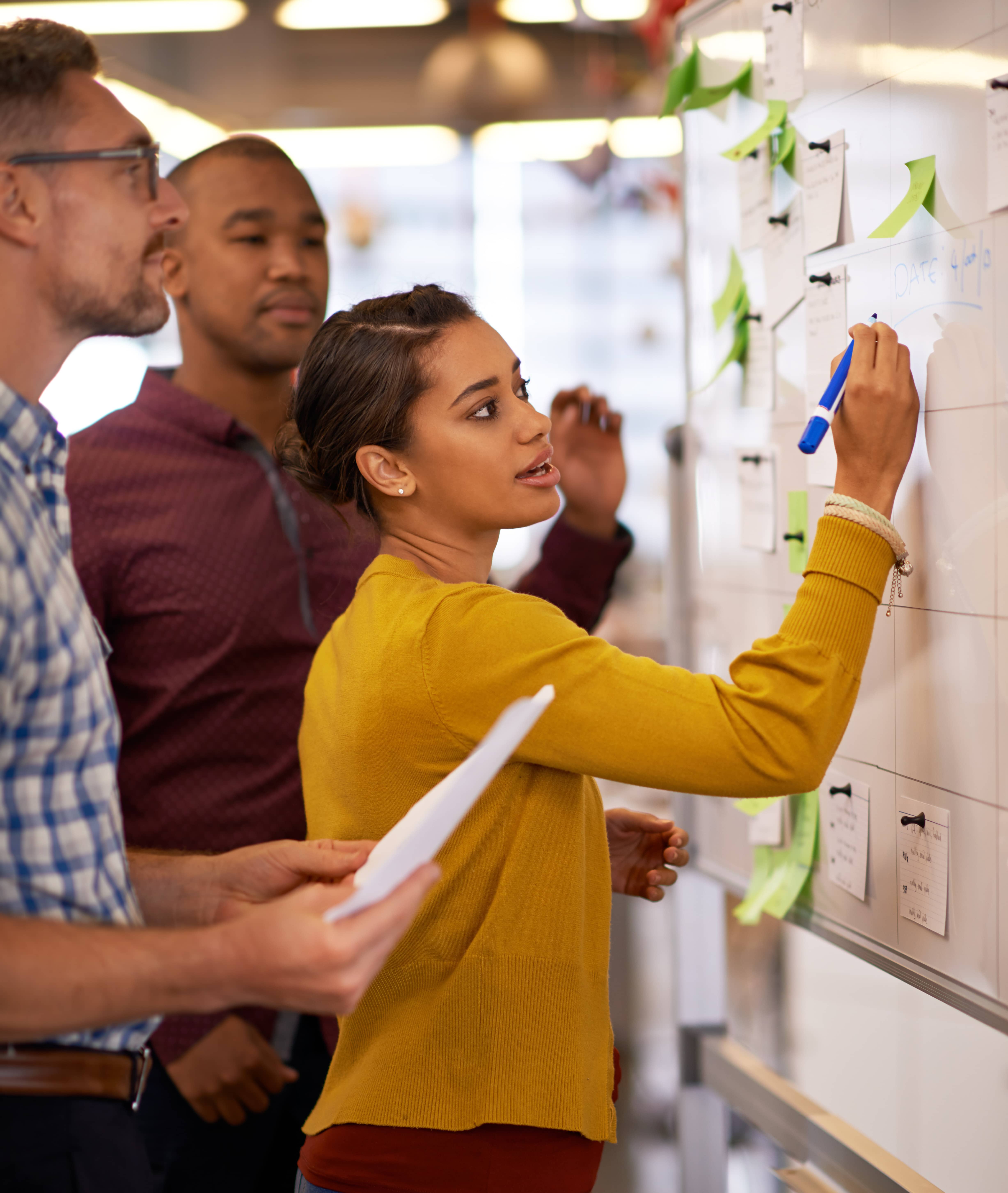 Woman in yellow sweater writes on whiteboard while two men watch and hold papers in office setting.