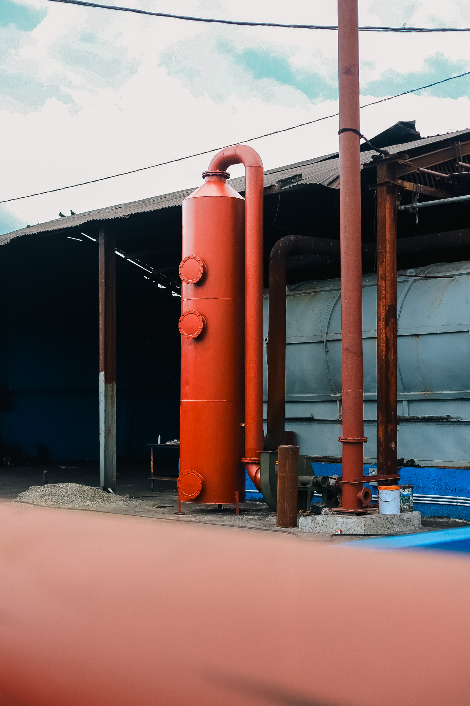 Industrial setting with a tall red cylindrical metal structure connected to pipes near a warehouse.
