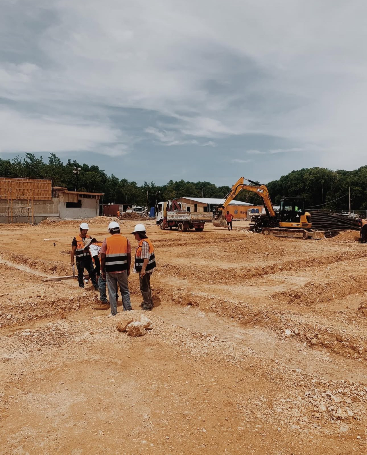 Construction site with workers in safety helmets and vests around excavated earth, with an excavator and trucks in the background.