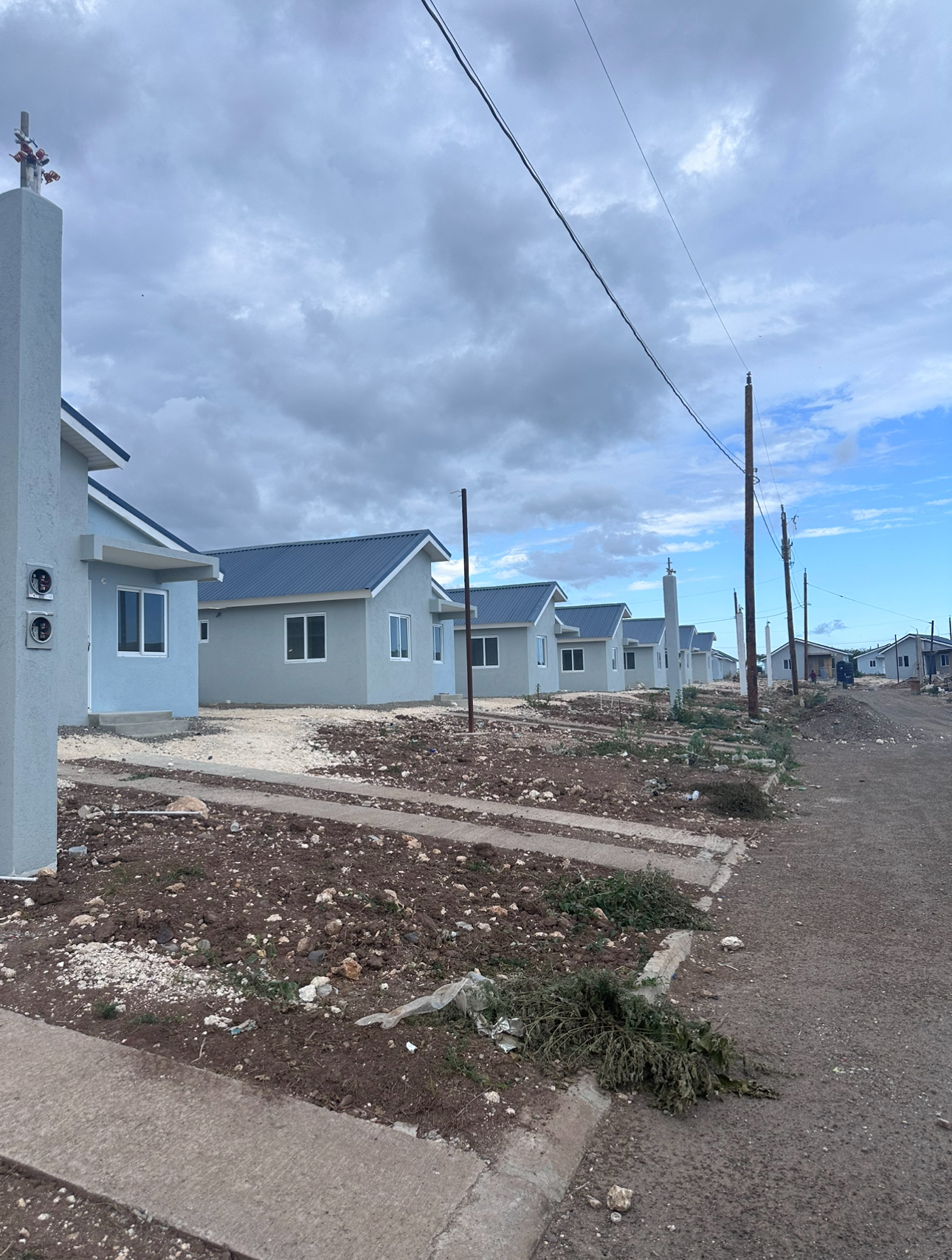 Row of newly built single-story houses with light blue walls and metal roofs along a dirt road under a cloudy sky.