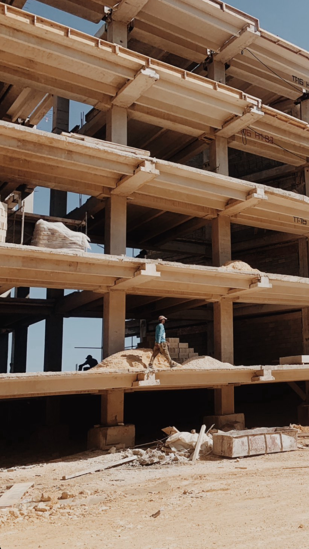 Construction site with an unfinished multi-story concrete building and a worker walking on sand inside.