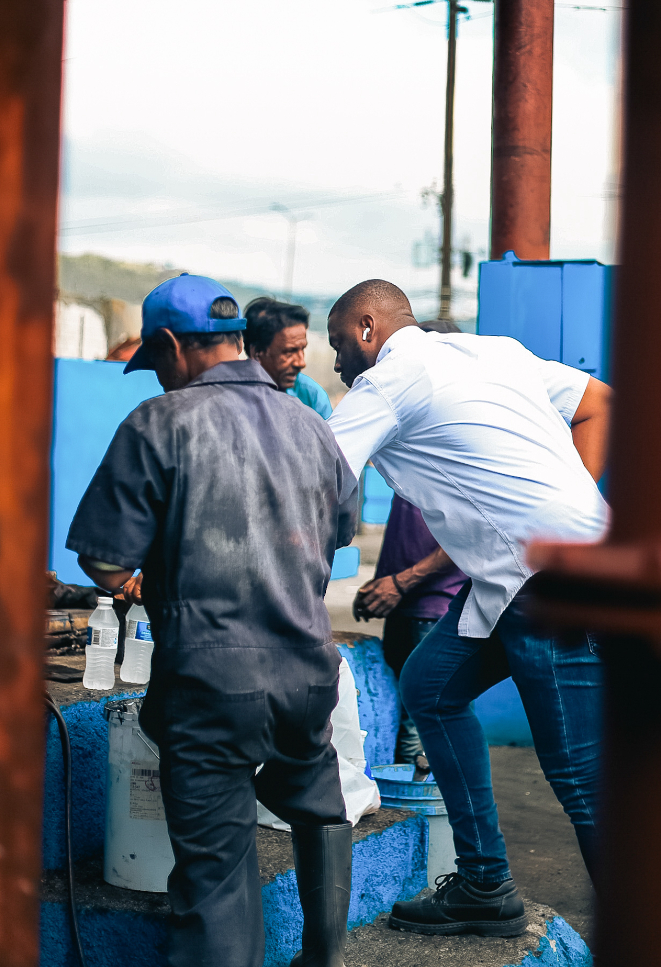 Three men interacting outdoors near blue-painted structures, one man in a white shirt bending forward.