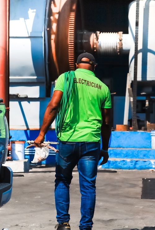 Man in green shirt labeled 'Electrician' carrying cable and tools walking outdoors near industrial equipment.