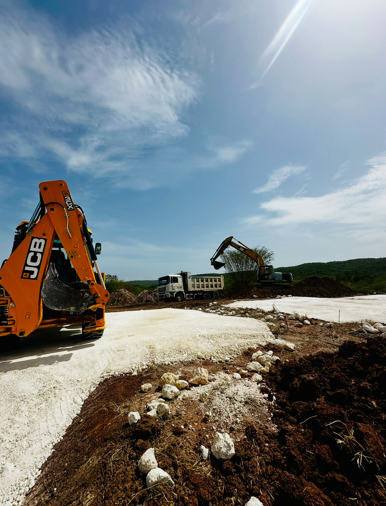 Construction site with a JCB excavator on the left, a CAT excavator in the background, and a dump truck, under a partly cloudy blue sky.