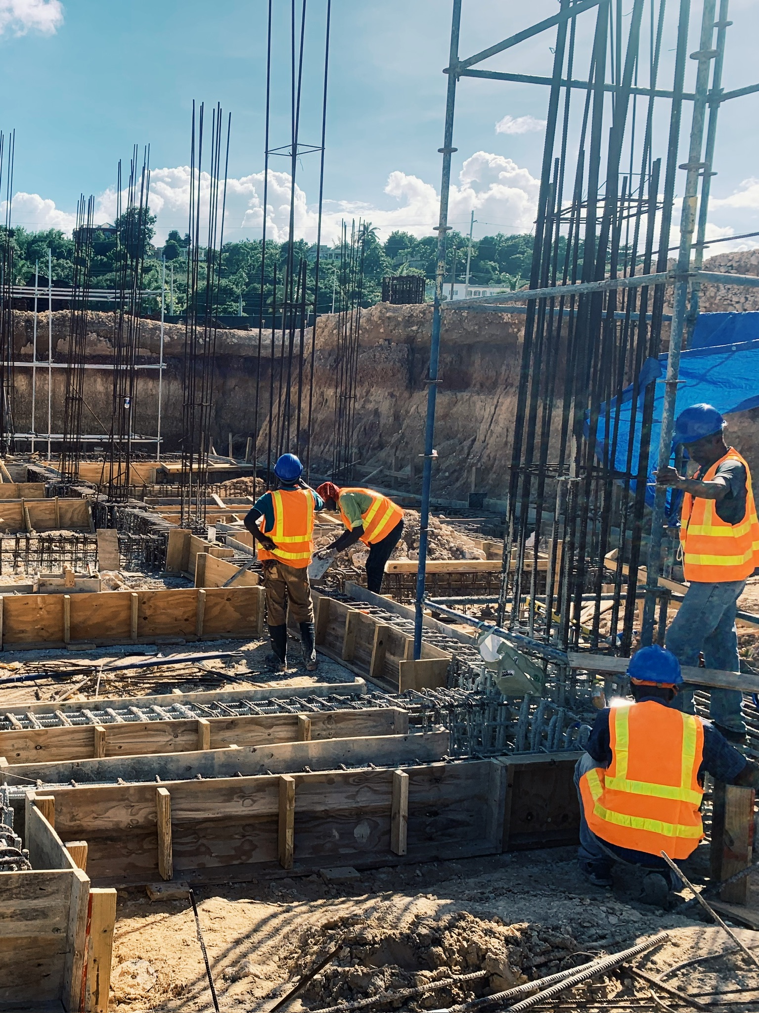 Construction site with four workers in orange safety vests and blue helmets working among steel reinforcement bars and wooden formwork under a clear sky.