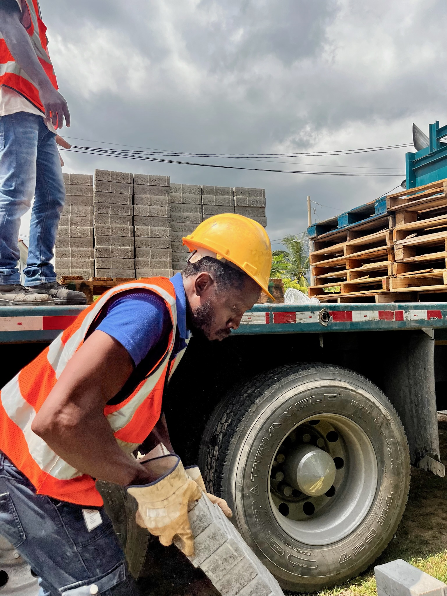 Construction worker wearing a yellow hard hat and orange safety vest lifting concrete bricks near a truck loaded with pallets and bricks.