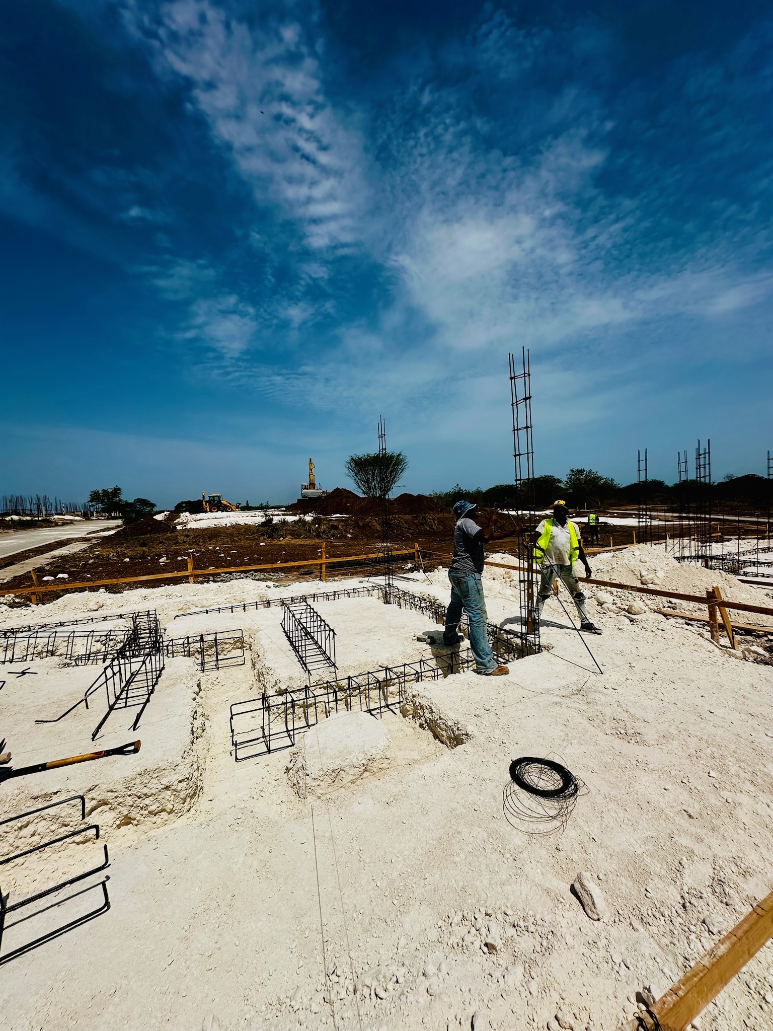 Two construction workers installing steel rebar framework on a sandy construction site under a blue sky with scattered clouds.