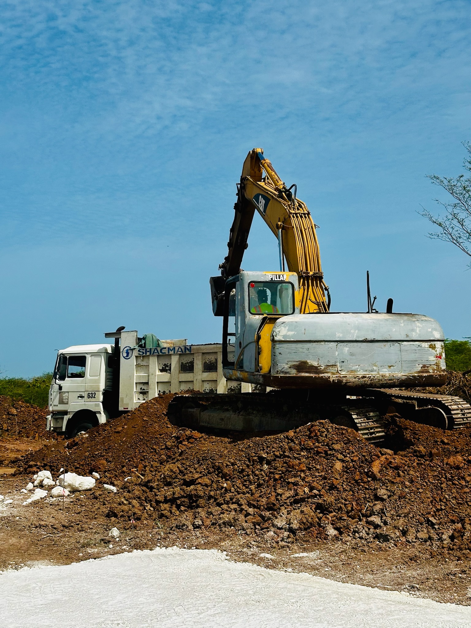 Yellow excavator loading dirt into a white Shacman dump truck on a construction site under a blue sky.
