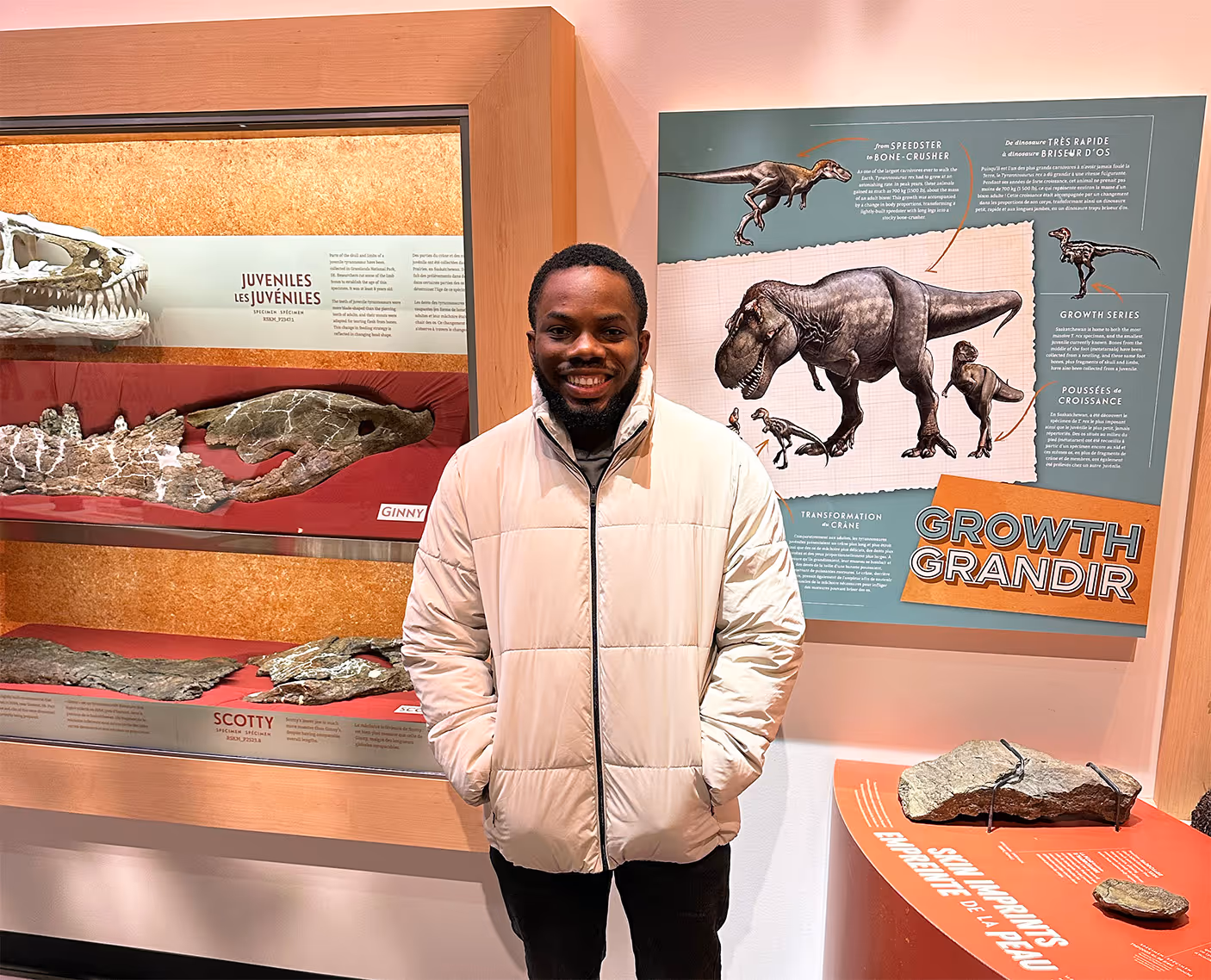 Smiling man in a light jacket standing inside a museum exhibit on dinosaur growth with fossils and educational posters.