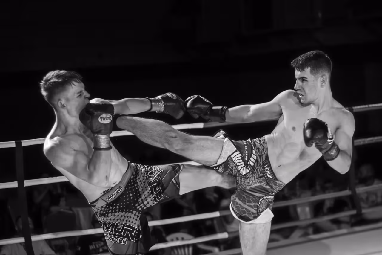 Zwei Kickboxer im Ring während eines Kampfes – einer trifft den anderen mit einem kraftvollen Tritt. Symbol für Durchsetzungsvermögen, Risiko und Strategie.