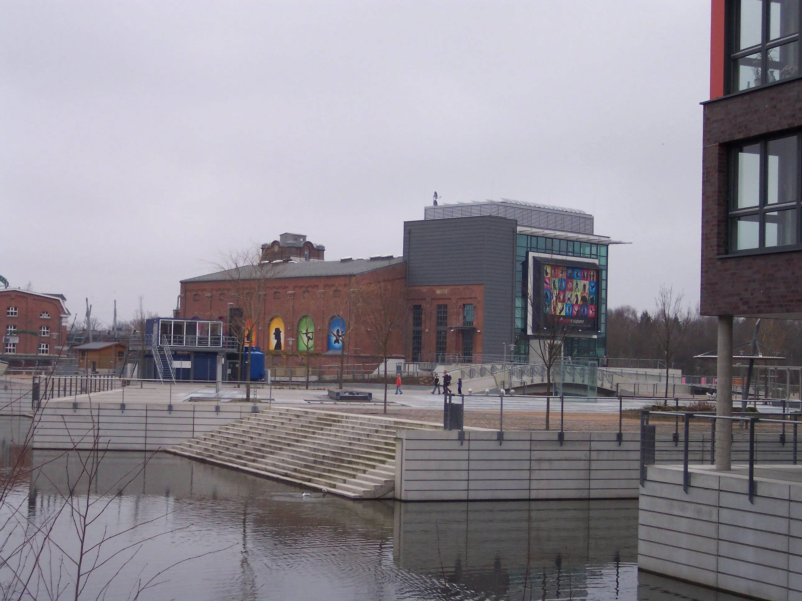 Ansicht des rock’n’popmuseums in Gronau mit moderner Glasfassade und historischer Backsteinhalle, davor eine breite Treppenanlage am Wasser.