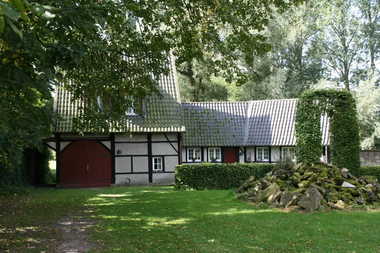 Historisches Fachwerkgebäude mit rotem Tor und grauem Ziegeldach in der Burganlage Nienborg in Heek, umgeben von Bäumen und einer Rasenfläche mit Steinansammlung.
