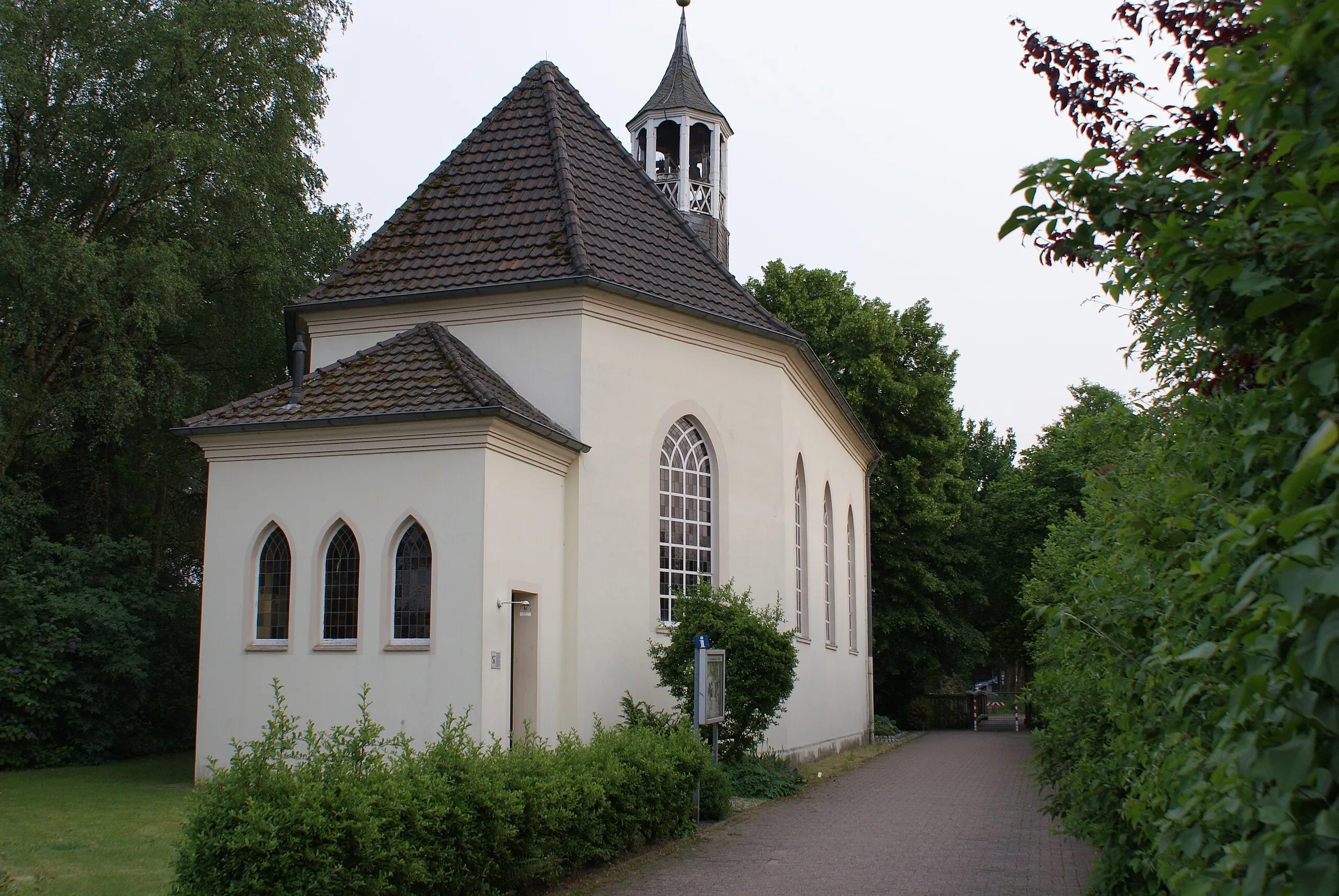 Evangelische Johanneskirche in Südlohn, weiß verputzter Bau mit hohen Spitzbogenfenstern und kleinem Glockenturm, umgeben von Bäumen und Grünflächen.