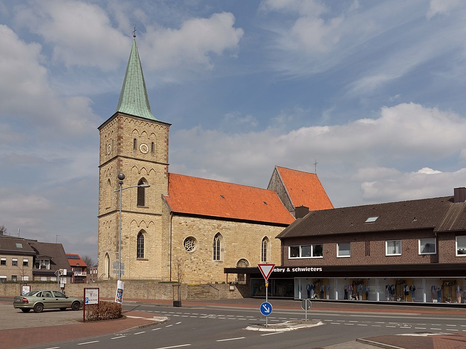 Historisches Fachwerkgebäude mit rotem Tor und grauem Ziegeldach in der Burganlage Nienborg in Heek, umgeben von Bäumen und einer Rasenfläche mit Steinansammlung.