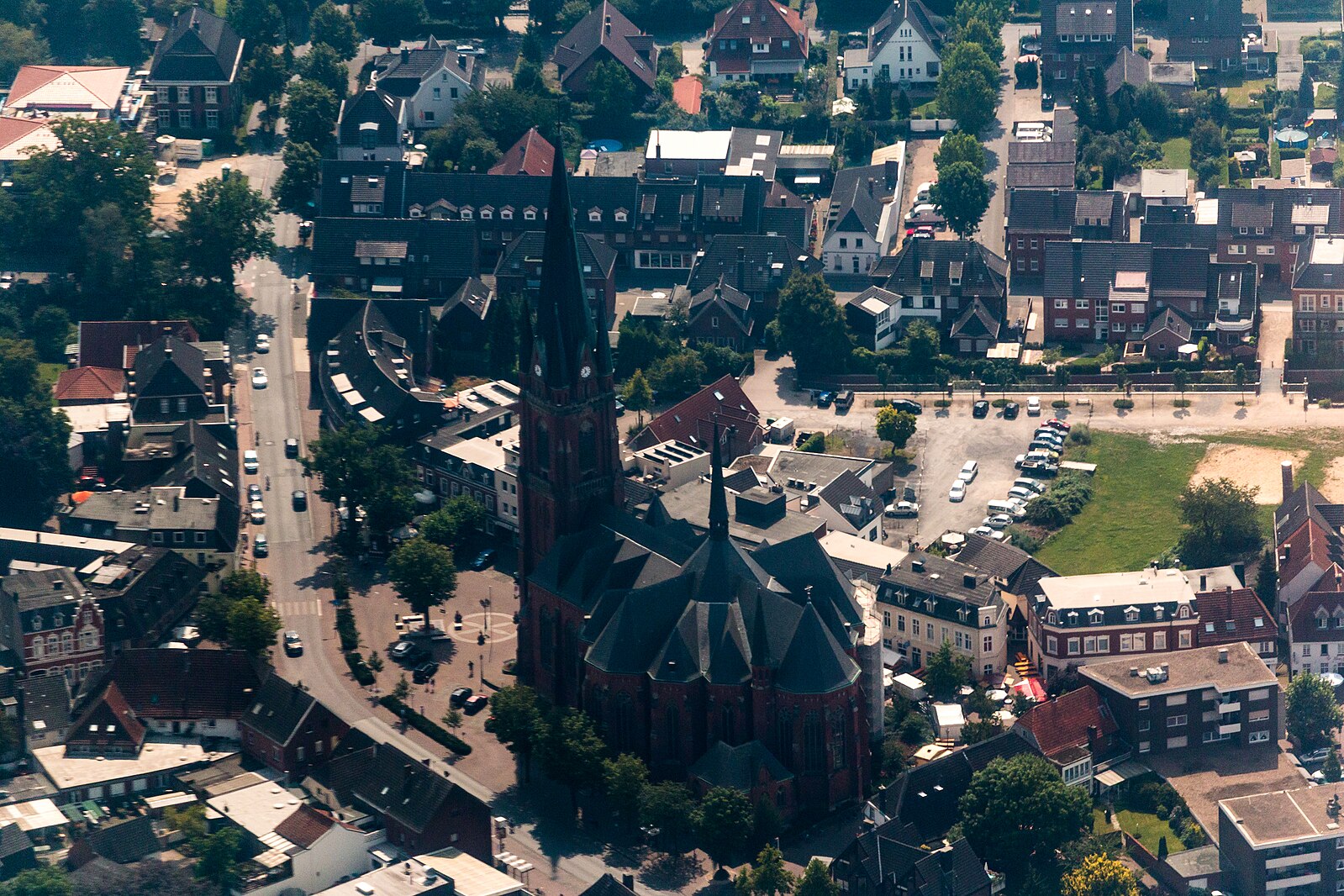 Luftaufnahme der St.-Gudula-Kirche in Rhede mit markantem roten Backsteinbau und hohem Turm, umgeben von Straßen, Wohnhäusern und Geschäften.