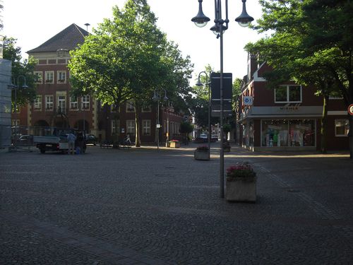 Blick auf den Marktplatz in Stadtlohn mit Kopfsteinpflaster, Blumenbeeten, Geschäften und historischen Backsteingebäuden im Abendlicht.